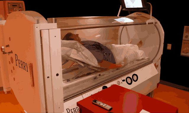 A person lying inside a Perry brand hyperbaric chamber with clear acrylic enclosure.