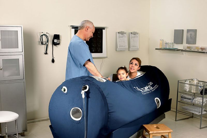 A doctor attends to a mother and child seated inside a Zuegma OxyHealth Hyperbaric Chamber in a clinical setting.