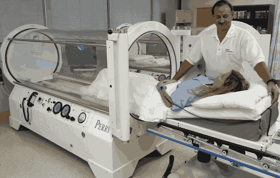 A patient lying on a bed is being guided into a Perry brand hyperbaric chamber by a medical professional — perry hyperbaric chamber.